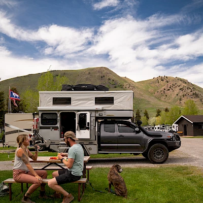 Two people are sitting at a picnic table near a camper truck with a dog sitting beside them, set against a scenic backdrop of mountains and houses.