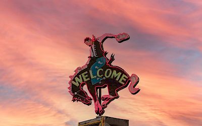 The image shows a sign featuring a cowboy on a bucking horse with the word "WELCOME" against a background of a colorful sunset.