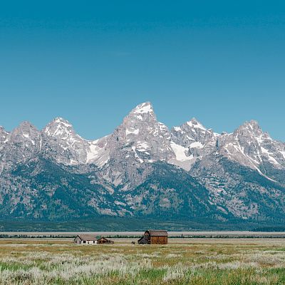 A scenic landscape featuring snow-capped mountains and green fields with a few rustic buildings in the foreground under a clear blue sky.