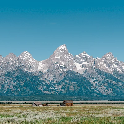 A scenic landscape featuring snow-capped mountains and green fields with a few rustic buildings in the foreground under a clear blue sky.