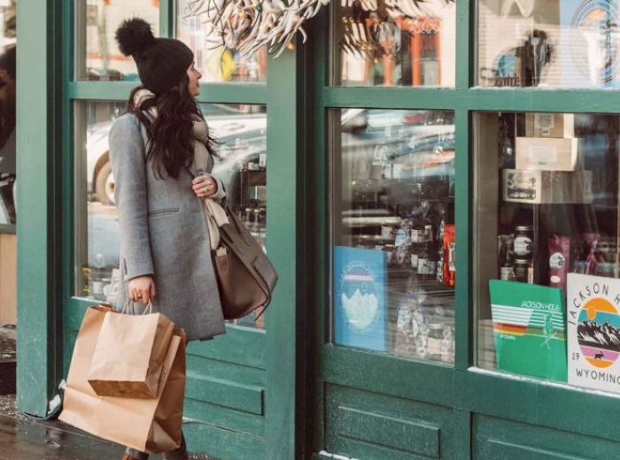 A person dressed in winter attire is standing outside a shop with green wooden frames, holding shopping bags and looking at the storefront.