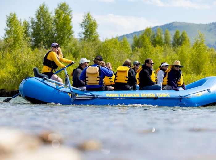 A group of people wearing life vests are rafting on a river in a blue inflatable raft, surrounded by trees and hills.
