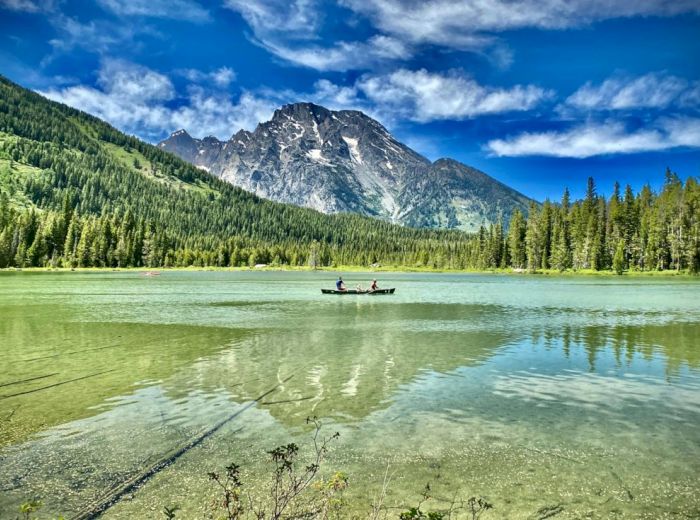 A serene mountain lake scene with clear water, a small boat in the center, and a backdrop of tall trees and rugged mountains against a partly cloudy sky.