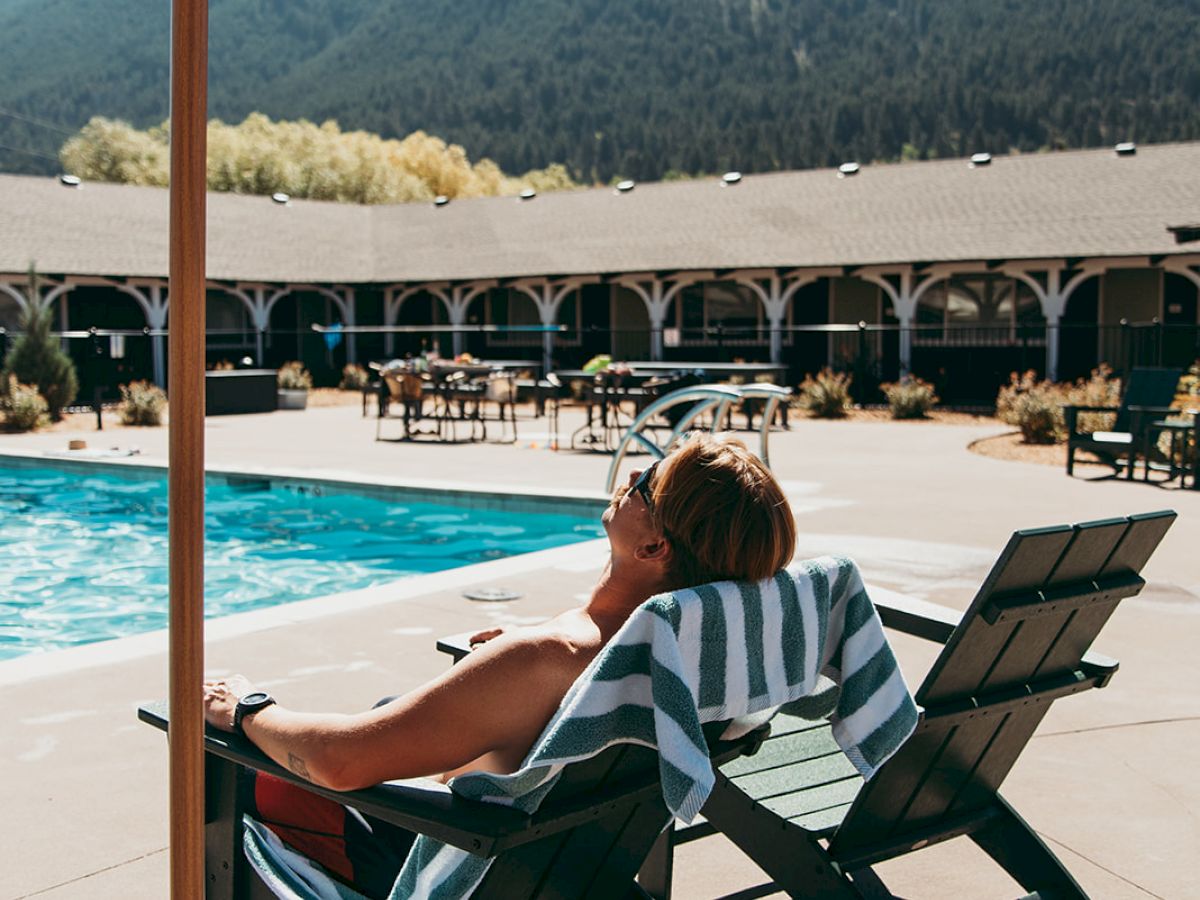 A person is lounging on a poolside chair under an umbrella, with mountains and a large building in the background on a sunny day.