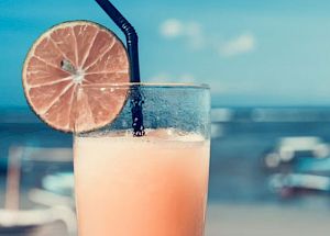 A hand holds a tall glass of a frothy drink with a black straw and a slice of lime, set against a beach scene with boats in the background.