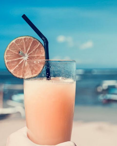 A hand holds a tall glass of a frothy drink with a black straw and a slice of lime, set against a beach scene with boats in the background.