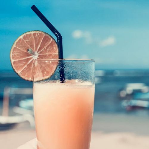A hand holds a tall glass of a frothy drink with a black straw and a slice of lime, set against a beach scene with boats in the background.